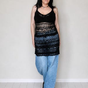 May include: A black sleeveless crochet top featuring a scoop neckline and an intricate openwork pattern throughout the body. The garment has a fitted bodice that transitions into a longer, semi-sheer design, paired with light blue denim jeans.