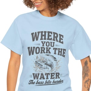May include: A light blue short-sleeve t-shirt featuring a dark gray graphic print. The text reads "WHERE YOU WORK THE WATER" above an illustration of a jumping bass fish, with the additional text "The bass bite harder" printed below.