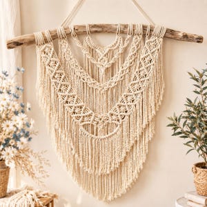 May include: A cream-coloured macrame wall hanging suspended from a natural wooden branch. The piece features intricate knotted patterns arranged in layered, V-shaped tiers, ending in long, flowing fringe that creates a pointed, symmetrical silhouette.