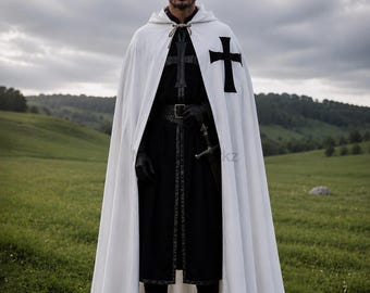Puede incluir: Una capa blanca con capucha que presenta un emblema de cruz negra en el pecho, sobre una túnica negra larga con un cinturón oscuro y guantes negros. El conjunto incluye una empuñadura de espada visible en la cintura y botas oscuras.