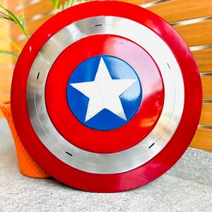 May include: A red, white, and blue shield with a silver ring. The shield features a white star on a blue circle in the centre. The outer ring is red, and the shield has a glossy finish. A decorative item.