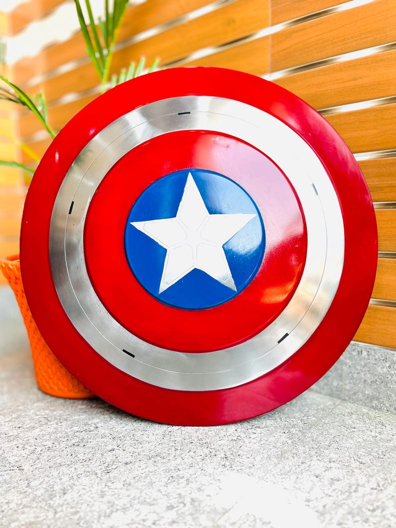May include: A red, white, and blue shield with a silver ring. The shield features a white star on a blue circle in the centre. The outer ring is red, and the shield has a glossy finish. A decorative item.