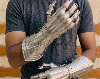 May include: A pair of articulated metal gauntlets, likely for historical reenactment or display. The gauntlets are silver-colored, with detailed finger and wrist protection. The text "Medieyworkz" is visible on the gauntlet. The gauntlets are designed to cover the entire hand and wrist.