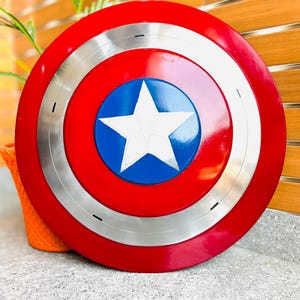 May include: A red, white, and blue shield with a silver ring. The shield has a white star on a blue circle in the centre. The outer ring is red, with a silver band separating it from the blue centre.