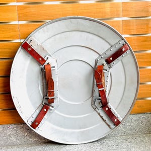 May include: A silver-coloured, round shield with a central circular indentation. It features two sets of riveted metal supports with red leather straps and buckles for securing the shield. The shield has a weathered appearance.