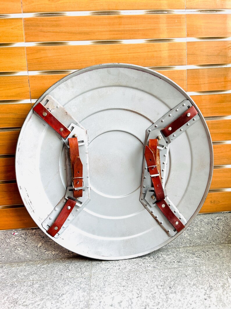 May include: A silver-coloured, round shield with a central circular indentation. It features two sets of riveted metal supports with red leather straps and buckles for securing the shield. The shield has a weathered appearance.