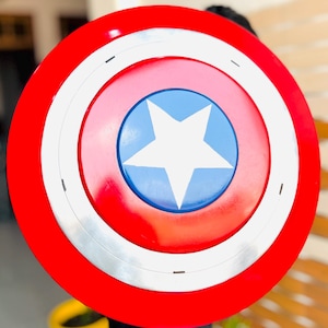 May include: A circular shield with a red border, a white band, and a blue central disc. A white star is in the middle. This shield is a replica of Captain America's shield.