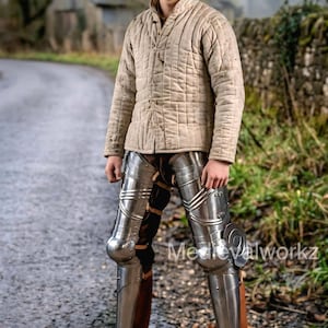 May include: A person wearing a tan quilted gambeson jacket and steel leg armor. The leg armor is highly polished and reflects the light. The person is standing on a paved road. The text "Medievalworkz" is visible on the image.