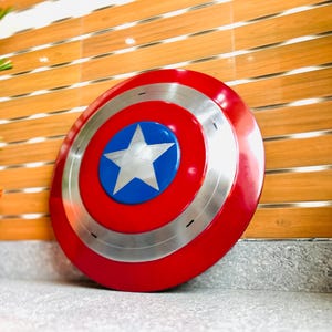 May include: A round shield in red, white, and blue, featuring a silver trim and a central white star. The design is iconic, resembling a superhero's shield, with a diameter of approximately 76 cm.