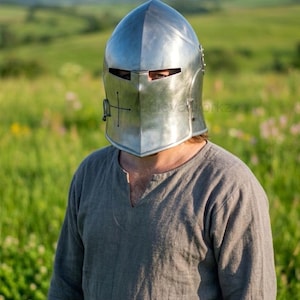 Puede incluir: Un yelmo medieval plateado y pulido con un diseño de cruz en la parte delantera. El yelmo tiene una visera con ranuras para los ojos y se lleva con una túnica gris. El fondo presenta un campo verde y un cielo azul.