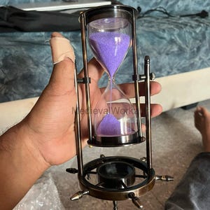 May include: An hourglass with a brass-coloured metal frame and a clear glass chamber filled with purple sand. The hourglass has a decorative base with a circular design and small, pointed accents. The text "MedievalWork" is visible on the glass.
