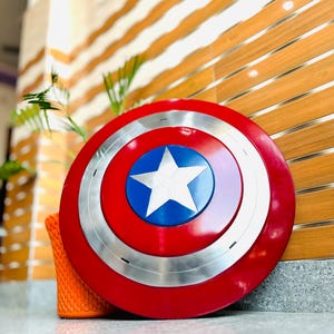 May include: A red, white, and blue shield with a star in the centre. The shield has a silver band and is leaning against a wooden wall. The shield is a replica of Captain America's shield.