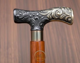 May include: A close-up of a walking cane with a decorative handle. The handle is a T-shape with ornate floral designs in silver and gold tones. The shaft is made of polished wood, and the cane has a brass collar.