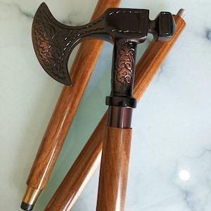 May include: A decorative walking cane with a bronze-colored axe head and handle. The axe head features an ornate floral design. The wooden shaft is a light brown color with brass accents. The cane is designed for support and style.