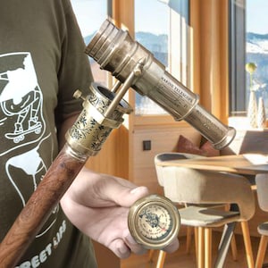 Puede incluir: Un telescopio marino de latón antiguo con el texto "MARINE TELESCOPE DOLLOND LONDON 1820". El telescopio está montado en un bastón de madera. Se sostiene una brújula de latón en la mano. El telescopio mide unos 46 cm de largo.