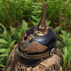 May include: A black and bronze Prussian Pickelhaube helmet. The helmet features a tall, pointed spike, decorative bronze emblems, and a leather visor. The helmet is displayed on a wooden stump against a backdrop of green foliage.