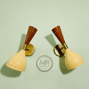 May include: Pair of vintage wall sconces with a mid-century modern design. Each sconce features a cream-colored, cone-shaped shade with small dot details, a brass-colored base, and a brown wooden top. The sconces are mounted on a light green wall.