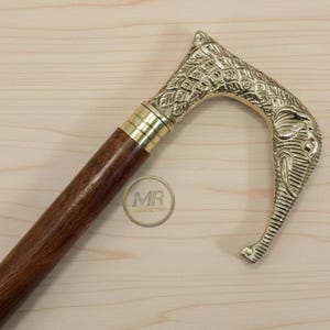 Può includere: Bastone da passeggio decorativo con manico a testa di elefante in ottone e fusto in legno marrone scuro. Il manico presenta dettagli intricati e una finitura dorata. Il bastone è progettato per supporto e stile.