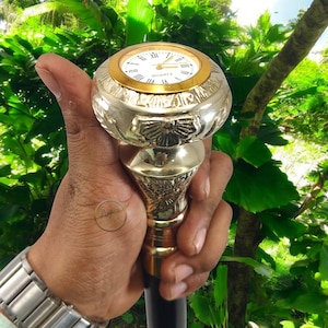 May include: A brass walking cane with a quartz watch embedded in the handle. The watch face is white with black Roman numerals and gold trim. The handle and shaft are intricately detailed.