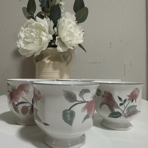May include: Four white ceramic planters with a floral design featuring pink flowers and green leaves. The planters have a pedestal base and a blue rim. A vase with white flowers and greenery is in the background.