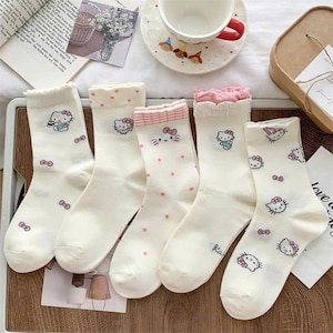 May include: Five pairs of white socks with various designs featuring a cartoon cat character. Some socks have pink bows, polka dots, or ruffled tops. The socks are arranged on a light-colored surface.