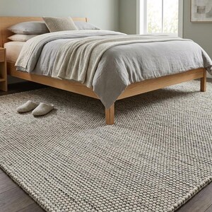 May include: A bedroom scene featuring a light wood bed frame with gray bedding and a cream-colored throw blanket. A textured, neutral-toned rug covers the floor, with a pair of slippers near the bed. A wooden nightstand with a lamp is visible.