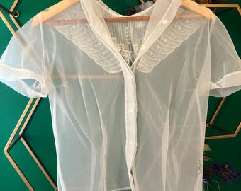 May include: A sheer white blouse with short sleeves and a button-up front. The blouse features a delicate lace collar and trim, and is hanging on a wooden hanger. The fabric appears lightweight and airy, suggesting a vintage or delicate style.