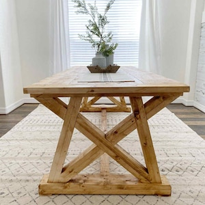 May include: A large, rectangular wooden dining table with a natural finish. The table features a trestle base with crossed supports and a thick tabletop. The table is set on a patterned rug, with a window and decorative items in the background.