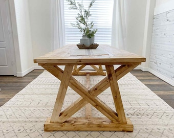May include: A large, rectangular wooden dining table with a natural finish. The table has a trestle base with crossed supports and a thick tabletop. The table is set on a patterned rug, with a window and decorative items in the background.