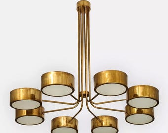 May include: A gold-toned chandelier with eight cylindrical light fixtures. Each fixture has a gold exterior and a white interior. The chandelier is suspended from the ceiling by multiple gold rods.