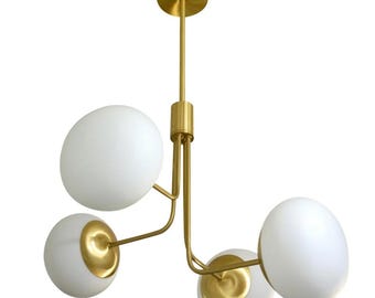 Könnte beinhalten: Eine moderne Deckenleuchte mit Messing-Finish und vier weißen Kugelschirmen. Die Leuchte hat einen zentralen Stiel und gebogene Arme, die die kugelförmigen Leuchten halten. Das Design ist minimalistisch und elegant, passend für verschiedene Einrichtungsstile.