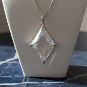 以下が含まれることがあります： 白いジュエリーバストに、大きな透明な菱形のペンダントが付いたシルバーのネックレスが飾られています。 ペンダントはファセットデザインです。 バストは濃い青色の表面にあります。