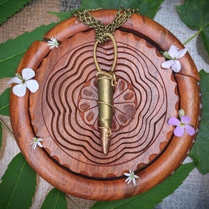 Puede incluir: Un collar con colgante de bala de latón con una cadena dorada. La bala está envuelta en alambre dorado y cuelga de una cadena. El collar descansa sobre un plato de madera tallada con pequeñas flores blancas y moradas y hojas verdes.