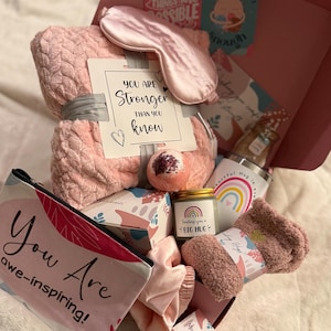 May include: A pink gift box filled with self-care items. Includes a blanket, eye mask, socks, bath bomb, candle, tumbler, and cosmetic bag. The cosmetic bag reads "You Are awe-inspiring!" and the candle says "Sending you a BIG HUG."
