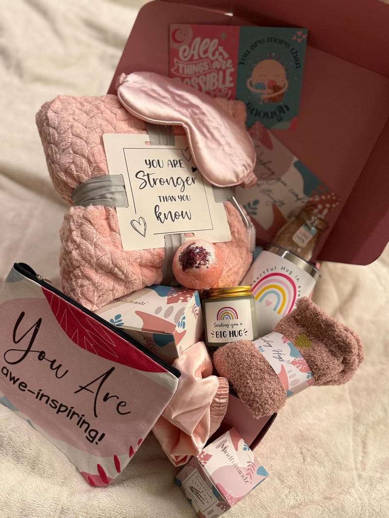 May include: A pink gift box filled with self-care items. Includes a blanket, eye mask, socks, bath bomb, candle, tumbler, and cosmetic bag. The cosmetic bag reads "You Are awe-inspiring!" and the candle says "Sending you a BIG HUG."