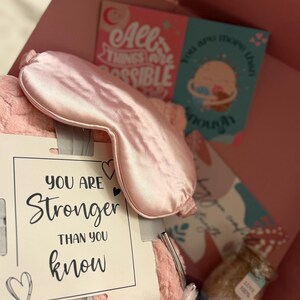 May include: A pink gift box filled with self-care items. Includes a satin eye mask, a plush pillow, a card with the text "You are stronger than you know", and other items. The box also contains a tumbler and a jar of bath salts.