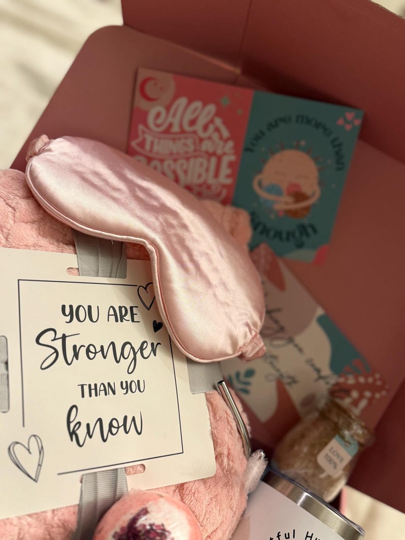May include: A pink gift box filled with self-care items. Includes a satin eye mask, a plush pillow, a card with the text "You are stronger than you know", and other items. The box also contains a tumbler and a jar of bath salts.