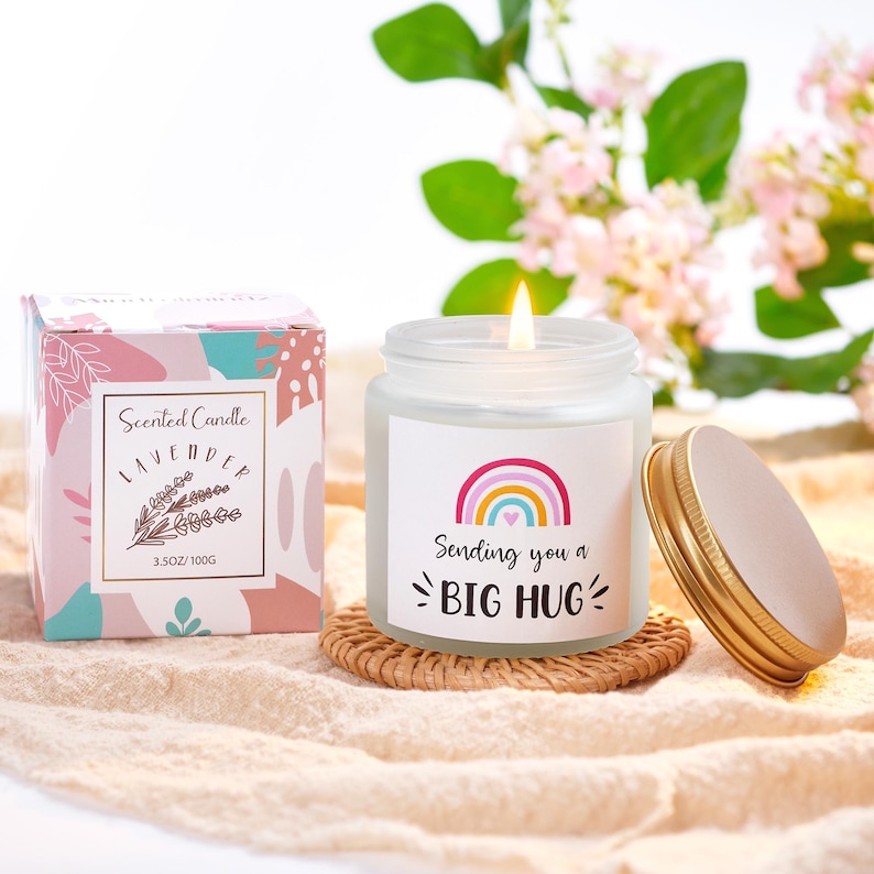 May include: A lit lavender-scented candle in a glass jar with a gold lid, next to a box. The candle has a label with a rainbow and the words "Sending you a BIG HUG." The box is pink and teal with the words "Scented Candle Lavender" and a sprig of lavender.