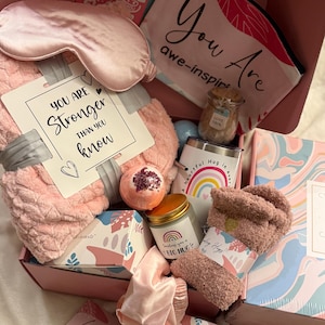 May include: A pink gift box filled with self-care items. Includes a sleep mask, blanket, socks, bath bomb, candle, tumbler, and cards with inspirational messages. The box is a thoughtful gift for relaxation and comfort.