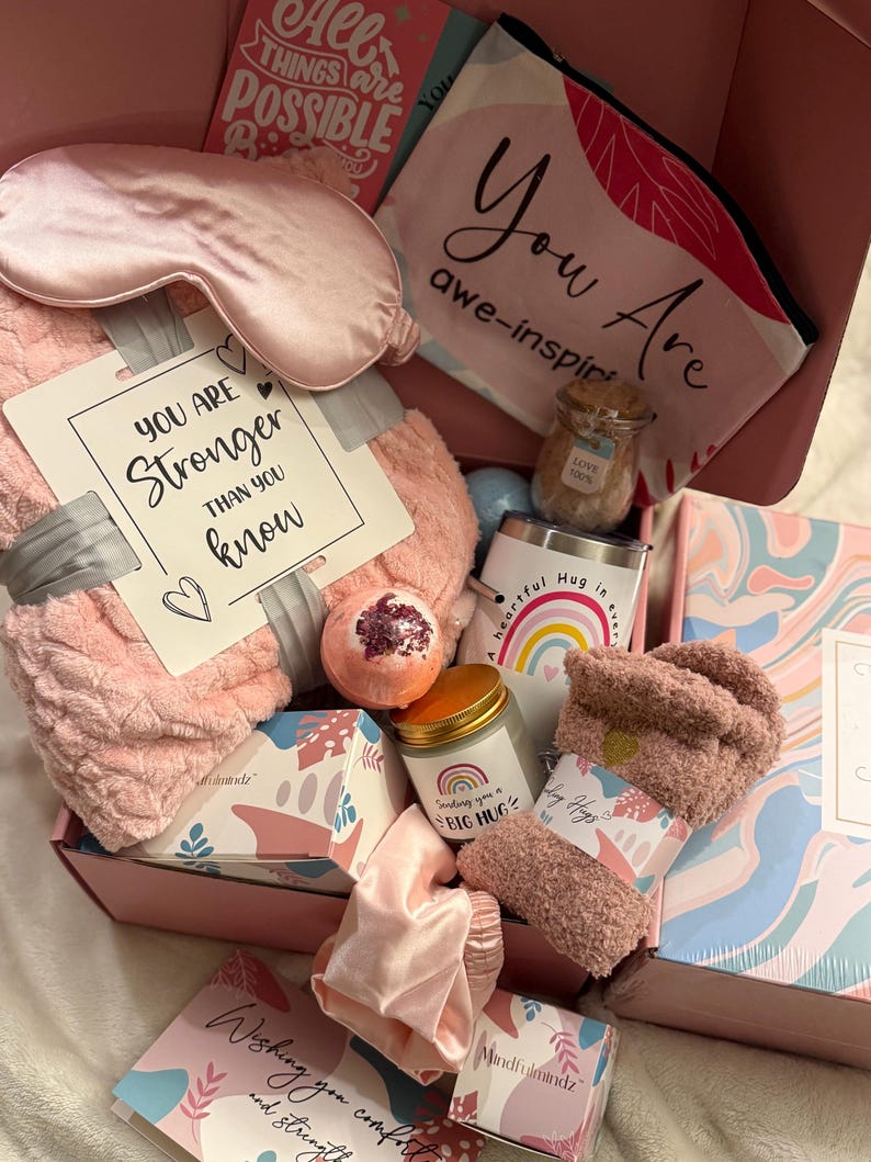 May include: A pink gift box filled with self-care items. Includes a sleep mask, blanket, socks, bath bomb, candle, tumbler, and cards with inspirational messages. The box is a thoughtful gift for relaxation and comfort.