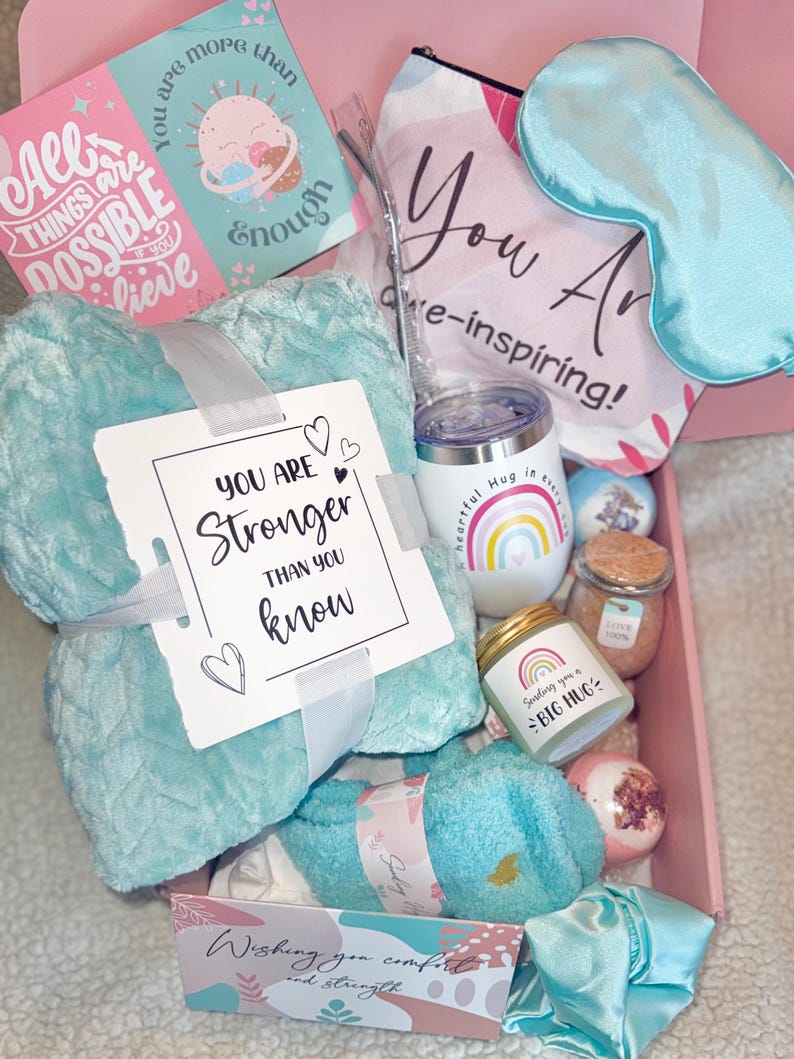 May include: A gift box filled with self-care items. Includes a teal blanket, eye mask, tumbler, candle, bath bombs, and cards with inspirational messages. The box is pink and contains items with the phrases "You are stronger than you know" and "Sending you a big hug."