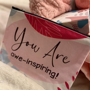 May include: A pink and white cosmetic bag with the words "You Are awe-inspiring!" in black script. The bag has a black zipper and is decorated with abstract floral designs. Other items in the image include a pink bath bomb and a fluffy pink spa headband.