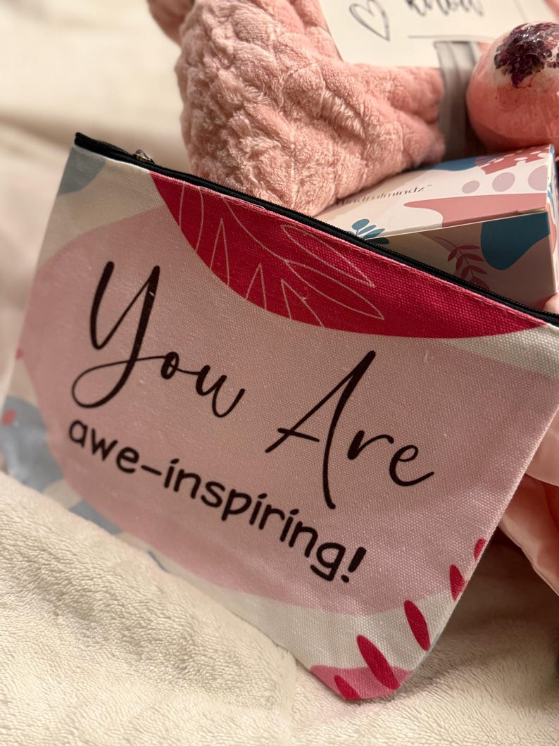 May include: A pink and white cosmetic bag with the words "You Are awe-inspiring!" in black script. The bag has a black zipper and is decorated with abstract floral designs. Other items in the image include a pink bath bomb and a fluffy pink spa headband.