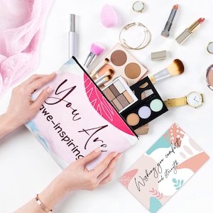 May include: A cosmetic bag with the words "You Are awe-inspiring" in a script font. The bag is open, revealing makeup brushes, eyeshadow palettes, and other beauty products. A card with the words "Wishing you comfort and strength" is also visible.