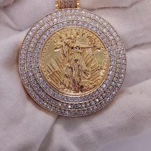 May include: A gold pendant featuring a detailed coin design with a central figure and the year 1998. The coin is surrounded by multiple rows of sparkling, clear gemstones. The bail is also encrusted with gemstones.