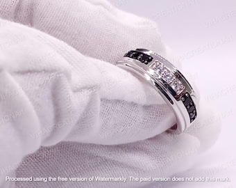 Puede incluir: Un anillo plateado con un diseño rectangular, con piedras negras y blancas. El anillo está sostenido por una mano enguantada, mostrando su superficie pulida y detalles intrincados. El diseño incluye filas de piedras negras y blancas.