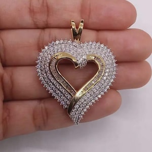 May include: A heart-shaped pendant crafted from yellow gold, featuring a central open heart design bordered by rows of sparkling white stones. The piece includes a gold bail at the top for attachment to a chain, showcasing a layered, textured appearance.