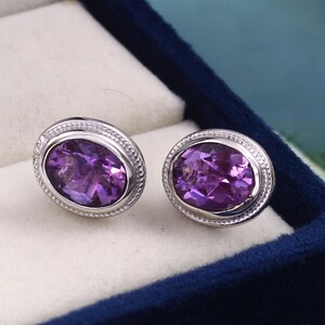 Könnte beinhalten: Ein Paar ovale Amethyst-Ohrringe. Jeder Ohrring hat einen leuchtend violetten Edelstein, der in einer silberfarbenen Lünette mit strukturiertem Rand gefasst ist. Die Ohrringe werden in einer dunkelblauen Schmuckschatulle mit weißem Innenraum präsentiert.