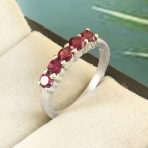 Könnte beinhalten: Ein silberfarbener Ring mit fünf runden, tief rosaroten Edelsteinen, die in einer Reihe oben auf der Ringschiene gefasst sind. Der Ring wird in einem neutralfarbenen Schmuckkästchen mit weichem, gepolstertem Innenfutter und dunkelbraunem Rand präsentiert.
