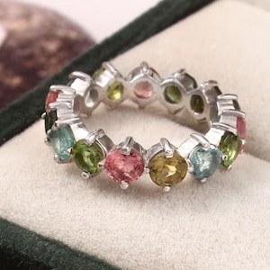 Könnte beinhalten: Ein silberner Ring mit einem Band aus bunten Edelsteinen. Der Ring zeigt eine Reihe von rund geschliffenen Edelsteinen in verschiedenen Farben, darunter Rosa, Grün, Blau und Gelb. Der Ring ist vor einem hellen Hintergrund platziert.